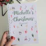 Hand holding a planner titled 'Michelle's Christmas Planner' with a cup of coffee and festive decorations in the background.