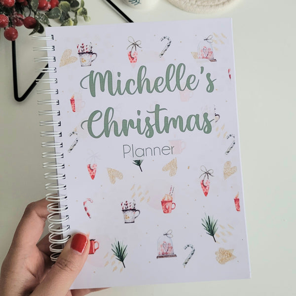 Hand holding a planner titled 'Michelle's Christmas Planner' with a cup of coffee and festive decorations in the background.