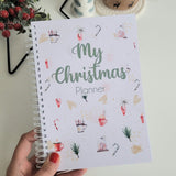 Hand holding a 'My Christmas Planner' with a cup of coffee and festive decorations on a white surface.